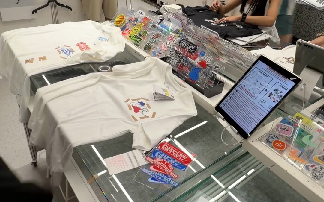 White tees laid on a Nike custom studio table in Seoul with sticker patches and a tablet